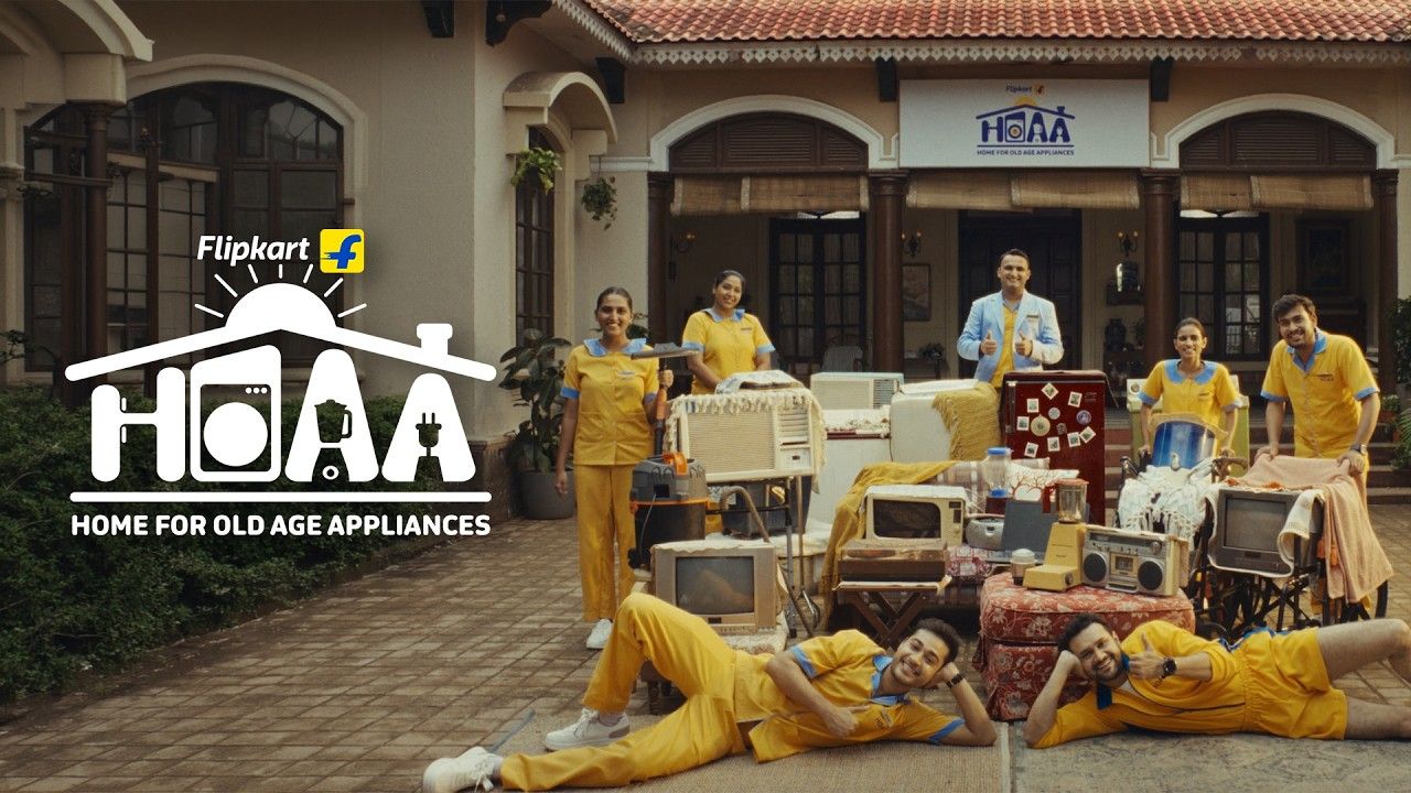 Old televisions, radios and appliances on a lawn in front of a house signed "HOAA" with staff in yellow uniforms, Flipkart Big Billion Days 2026